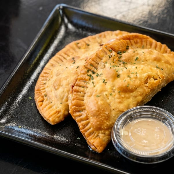 Two golden fried empanadas sprinkled with herbs sit on a black plate beside a small cup of creamy dipping sauce.