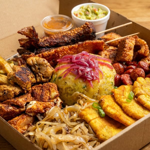 Open cardboard box filled with assorted grilled meats, fries, rice, vegetables, fried sides, dipping sauces, and pickled onions.