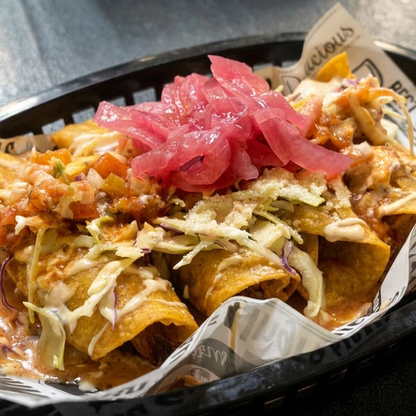 Crispy rolled tacos topped with pink pickled onions, shredded cabbage, creamy sauce, and salsa in a lined basket.